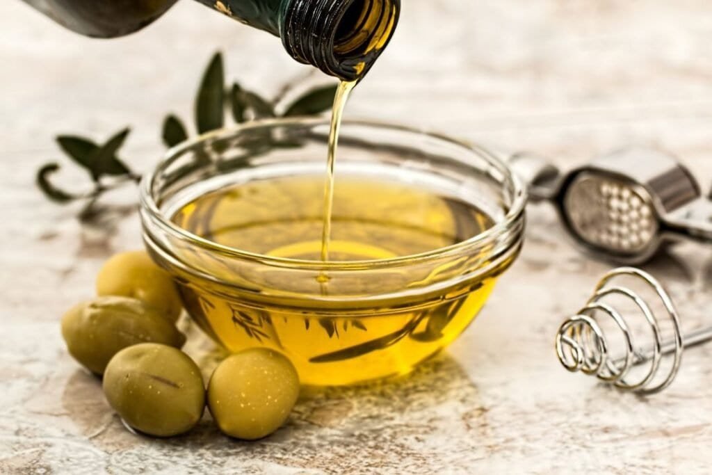 Pure olive oil being poured into a glass bowl with green olives and kitchen tools, emphasizing natural, healthy, and holistic home cooking. Fresh ingredients and eco-friendly practices support a holistic lifestyle for your home.